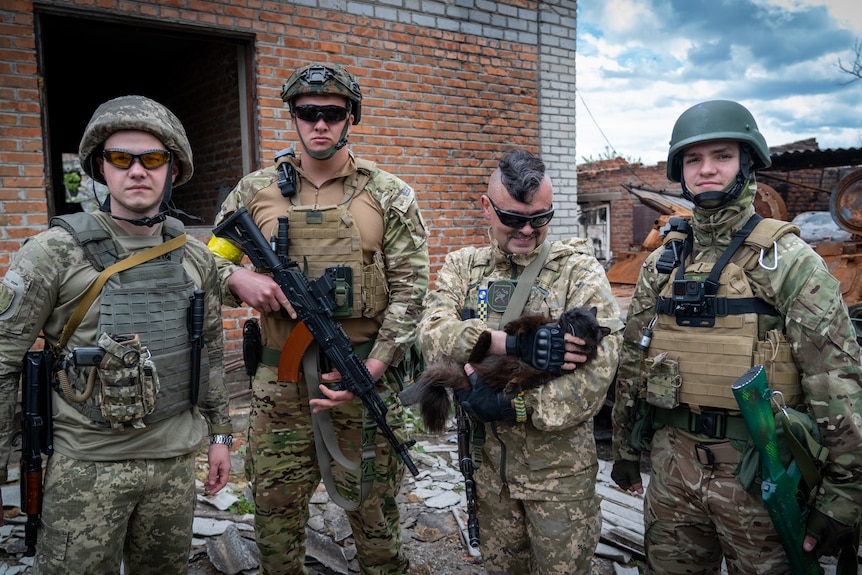 Four soldiers in army fatigues stand outside a brick home, one with a mohawk smiles adoringly at a black cat in his arms