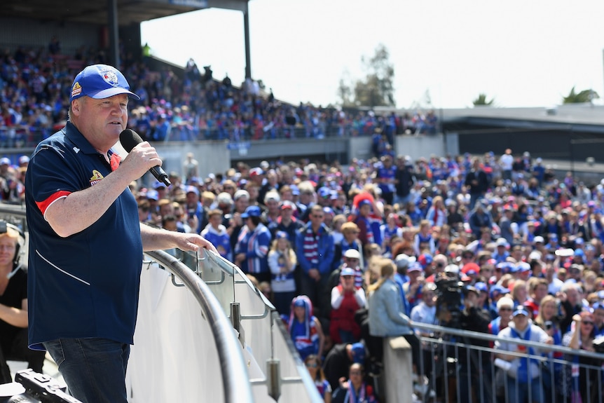 A man in a football polo and cap speaks to a crowd from a stage