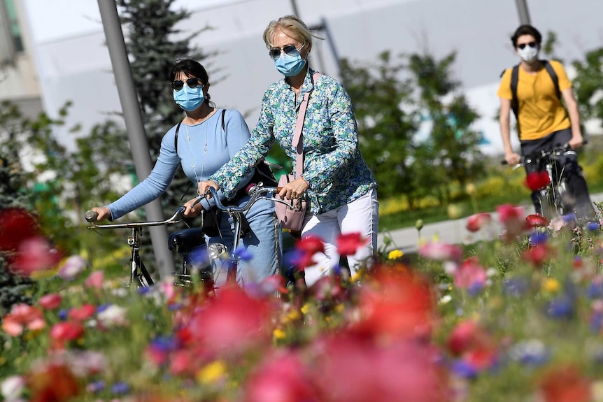 Women in masks walk with their bikes next to bright flowers