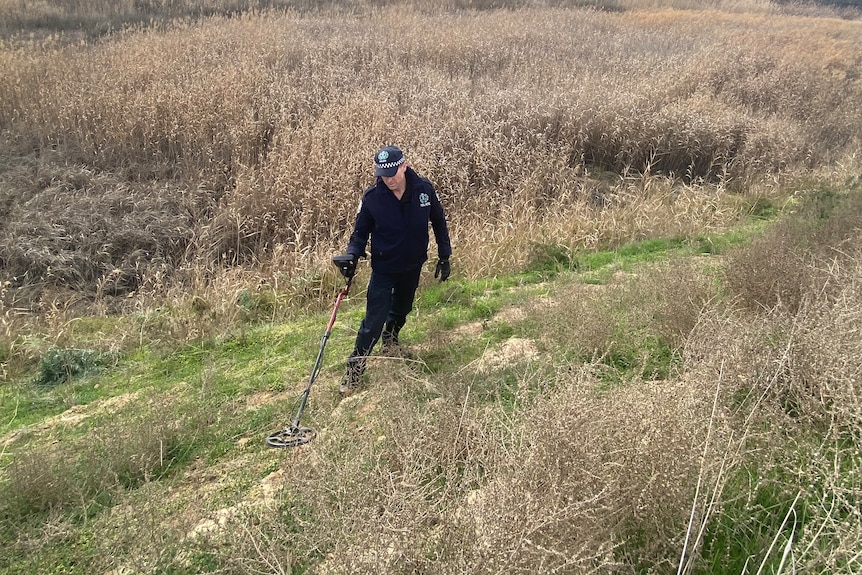 A police officer searches with a metal detector among grass in a rural area