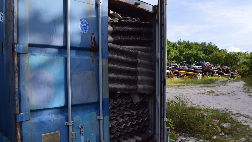 A blue shipping container's door is open, and sheets of asbestos are packed to the top.