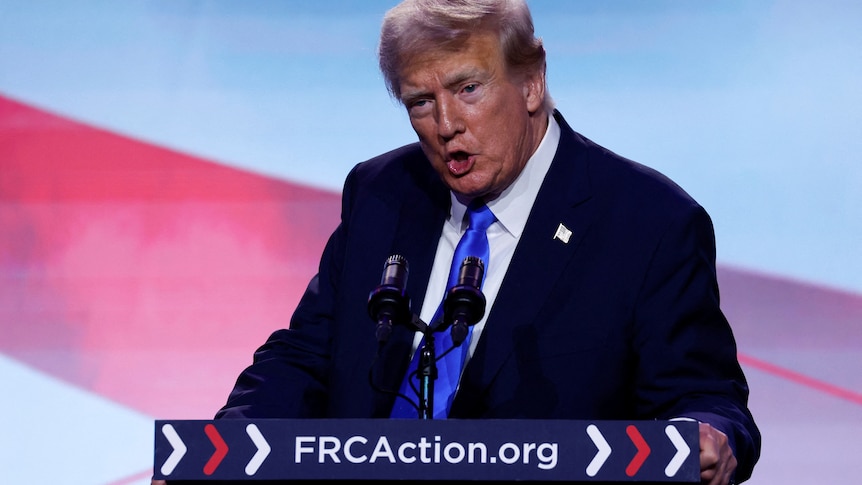 Donald Trump speaks at a podium wearing a dark suit and blue tie