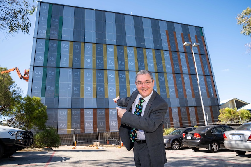 A man in a suit grins and points behind him at a building-sized periodic table on a university campus.