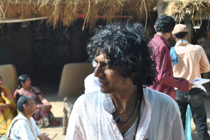 Girish looks to the side, his shirt open slightly showing a number of necklaces. Behind him some men and women sit on the ground