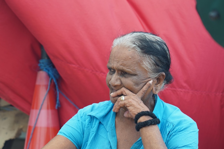 An older woman in a blue shirt rests her chin on her hand, sitting in front of a red tent