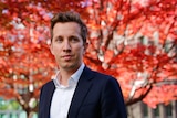 A young, Caucasian man in a suit is pictured, with a blossomed tree with bright red leaves in the background.