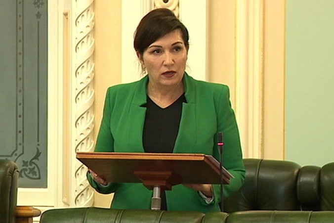 Woman standing at a lectern with a green jacket, black top and she looks serious and is not smiling.