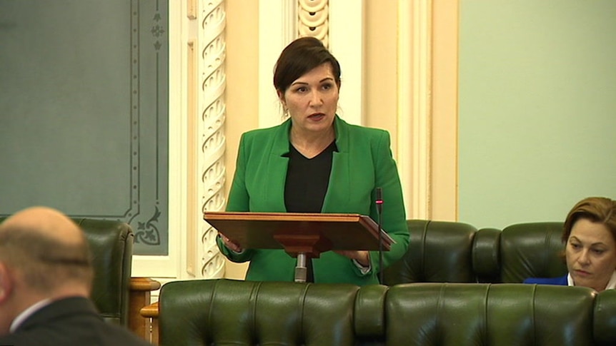 Woman standing at a lectern with a green jacket, black top and she looks serious and is not smiling.