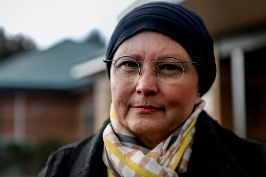 Woman in black coat and scarf wearing glasses stands outside aged care home beneath darkened skies