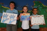 Three primary school children stand against a mural-painted wall holding Aboriginal language teaching materials.