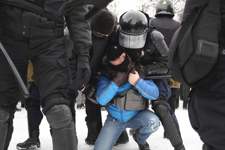 A police officer in St Petersburg grapples a protester, who is on his knees, from behind with his arms around his shoulders.