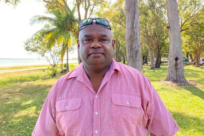 An Aboriginal man wearing a pink shirt and sunglasses atop his head stands in front of a tropical background and beach