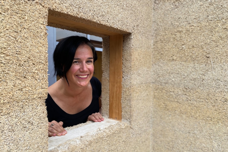 A black haired woman peaks through the window of a house made from rough, brown 'hempcrete'