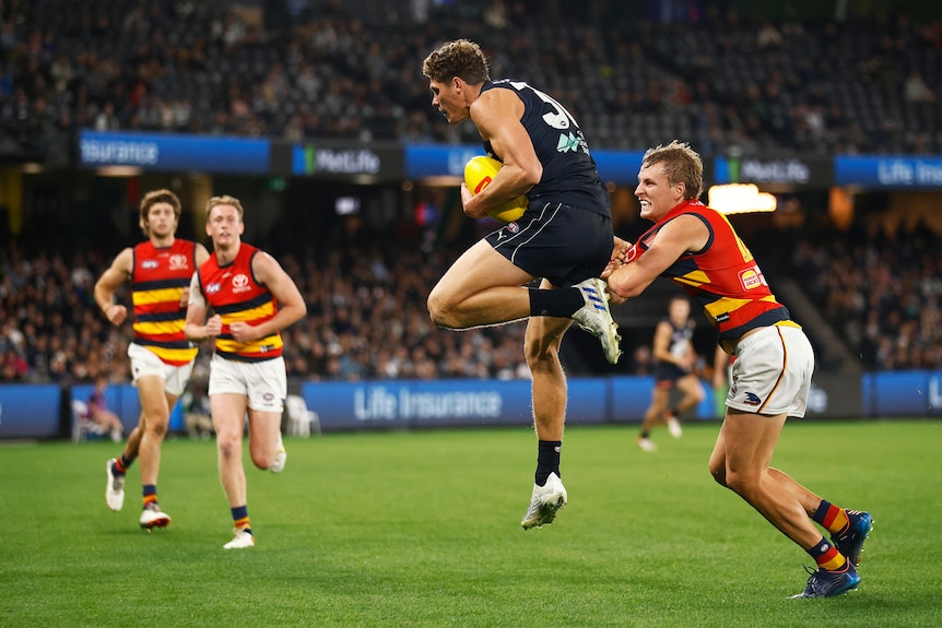 A Carlton forward holds the ball in to his chest while mid-air, as an Adelaide defender is a step behind. 