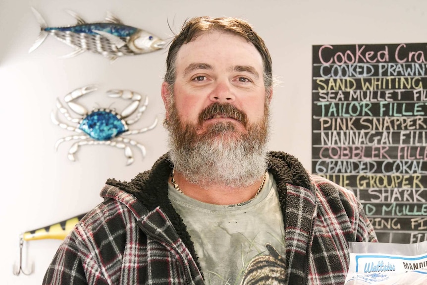 Brendon Watts standing in his fish shop holding a dead blue-swimmer crab