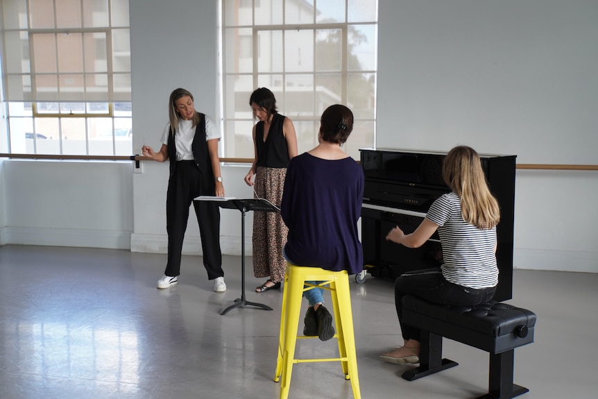 Four women in a room with a piano look at sheet music on a music stand.