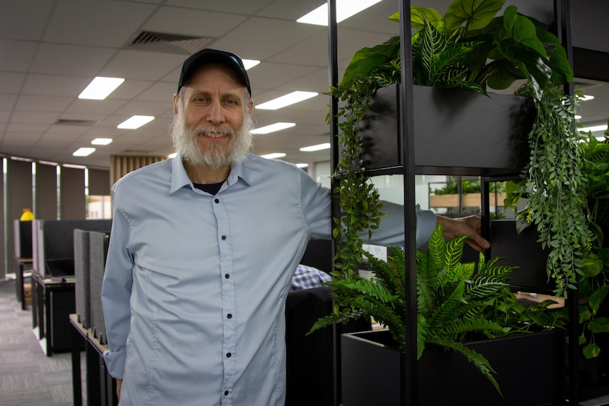 An older man with a grey beard and light blue shirt stands in an office, next to some house plants.