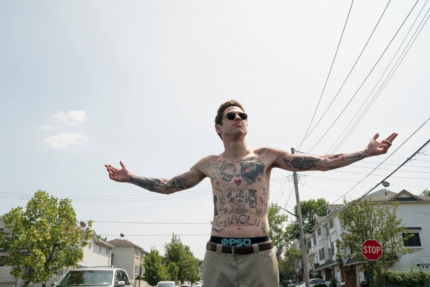 A shirtless man with tattoo covered upper body and sunglasses stands with arms spread on suburban street on sunny day.