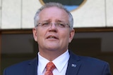 Scott Morrison, wearing a navy suit and orange tie, stands at a lectern with two microphones in front of two Australian flags.