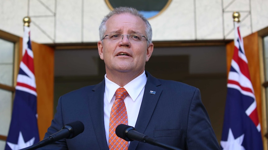 Scott Morrison, wearing a navy suit and orange tie, stands at a lectern with two microphones in front of two Australian flags.
