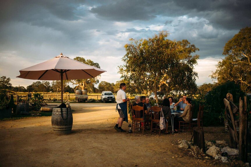 Seven people eat and drink from a wooden table outside next to leafy gum trees under a sunset sky while a waiter talks to them.