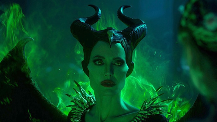 Angelina Jolie with black horns and wings is surrounded by bright green visual effects, and faces blonde figure with long hair.