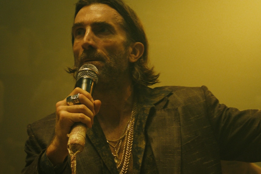 A film still of Sharlto Copley. He is holding a microphone to his mouth, the other arm outstretched to his left.