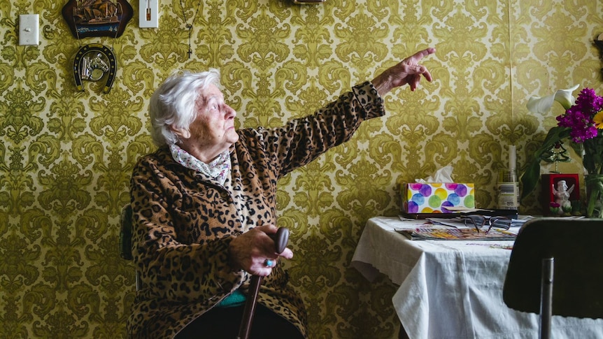 Valeria Miller sits holding a cane, she points at paintings above a table