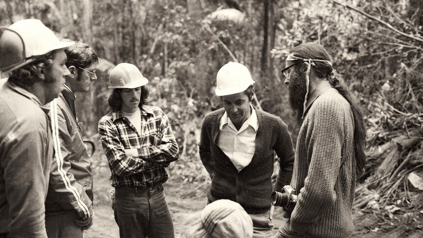 A black and white photo of a man with long hair and a camera talking to men in helmets in a forest.