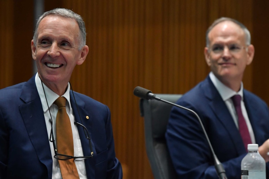 Two grey-haired men wearing blue suits and ties smile