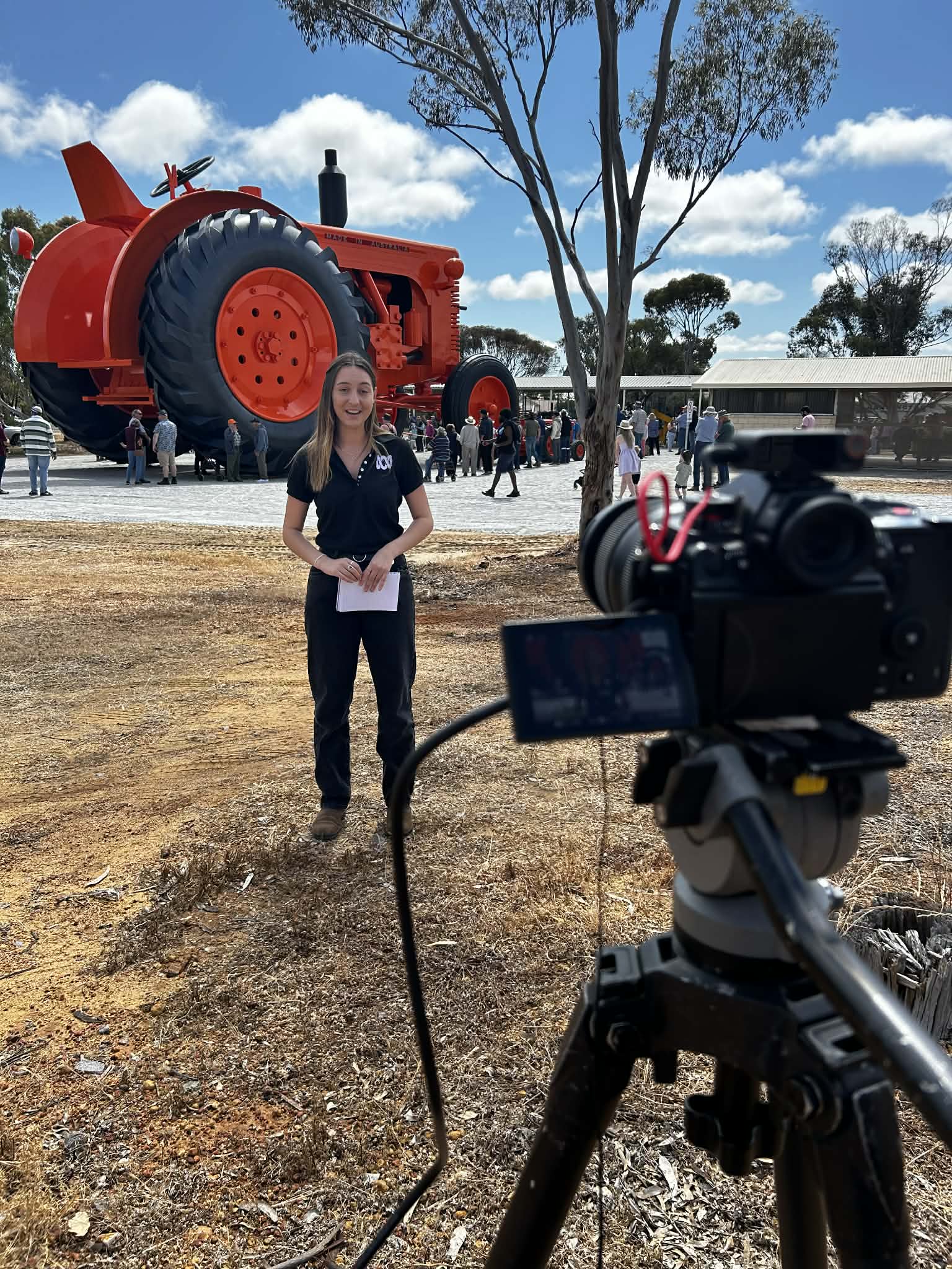 A reporter stands in front of a large orange tractor statue speaking to a camera on a tripod