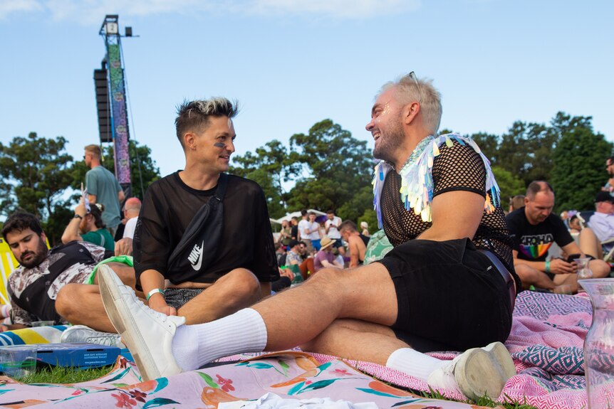 Two people with fashionable haircuts smile at each other as they sit on a rug laid out in a city park.