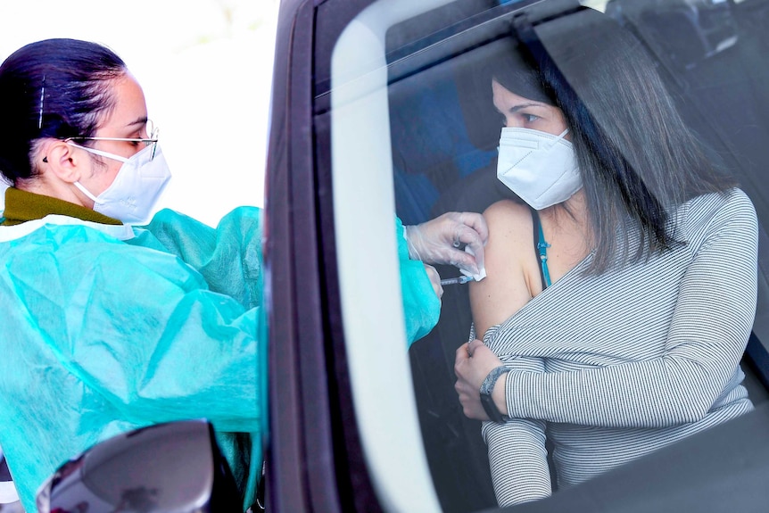 A woman in a face mask pulls her top down from her shoulder in a car while a nurse injects her through the window