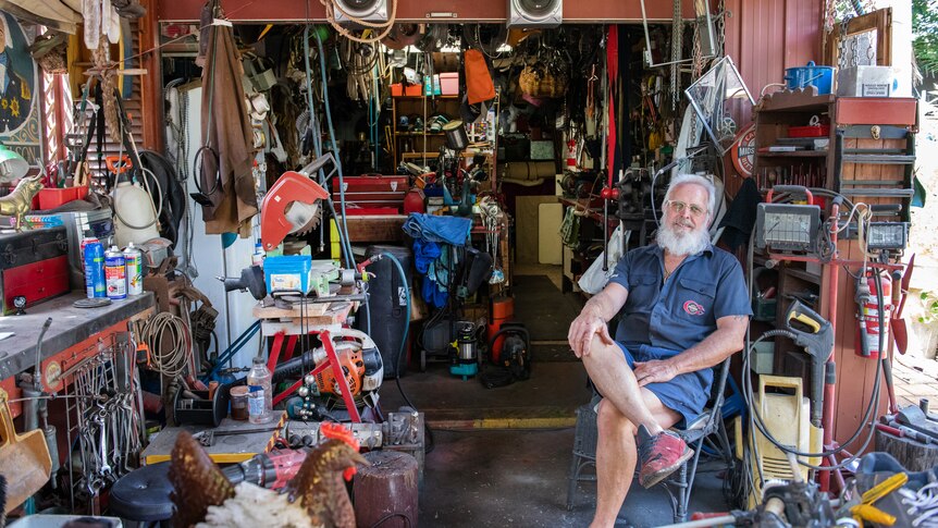 Wide shot of a man with a white beard seated on the right of his shed, which is well organised but full of stuff.