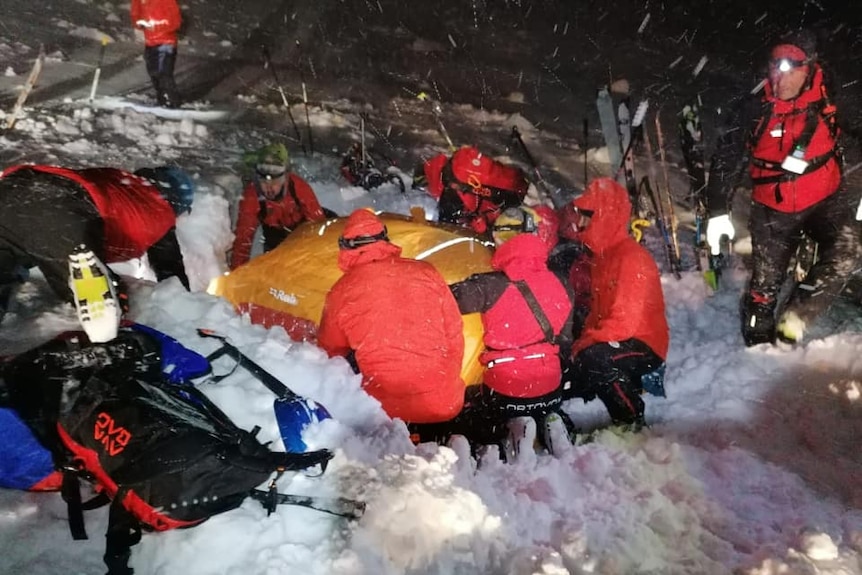 A group of people in bright red jackets dig under an orange tarp over the snow.