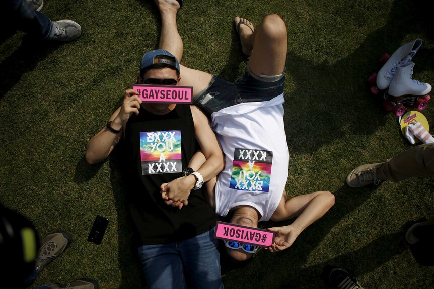 Two young men lying on the grass holding hands, with signs over their faces reading #GaySeoul