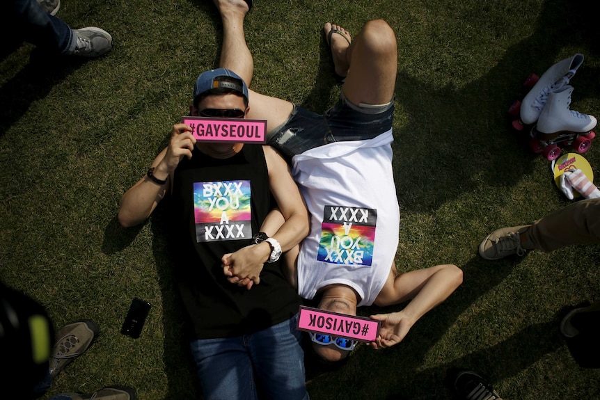 Two young men lying on the grass holding hands, with signs over their faces reading #GaySeoul