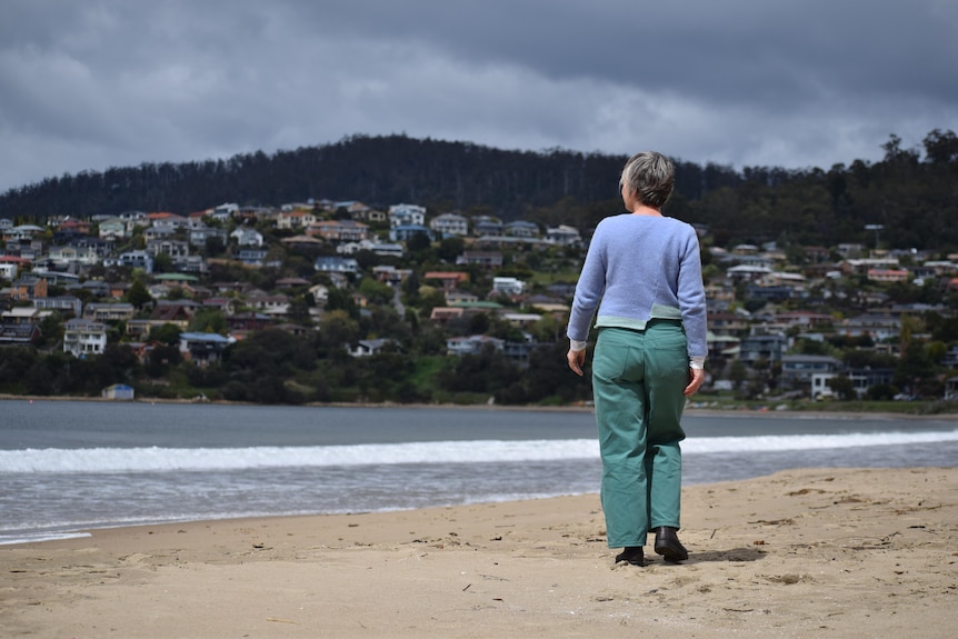 A woman walking away from camera on beach, houses on hills in background. She wears blue jumper and green pants. 