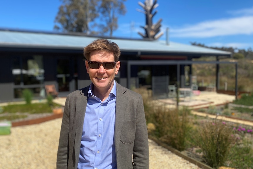 a man in sunglasses in a suit smiling in front of a country home with solar panels