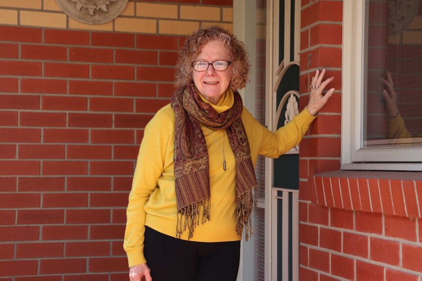 A woman wearing glasses and a patterned maroon scarf smiles while standing before the front door of a brick house.