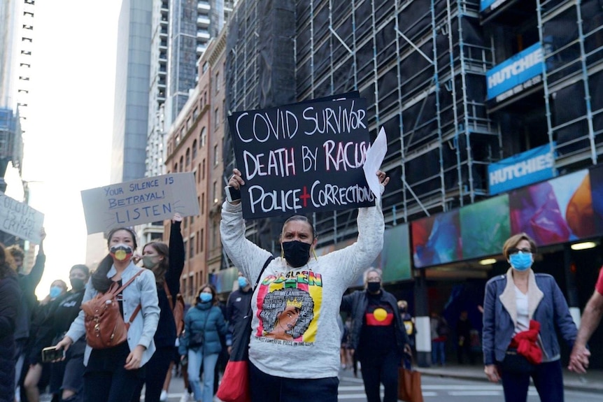 A woman is seen holding a sign reading COVID SURVIVOR DEATH BY RACISM
