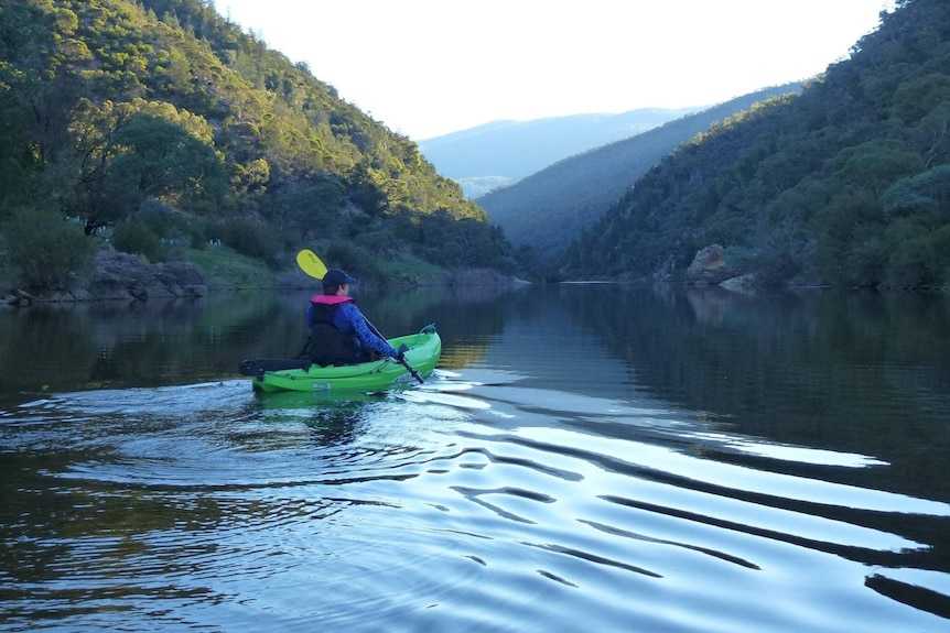 A woman paddles on a kayak along a river on a sunny day.