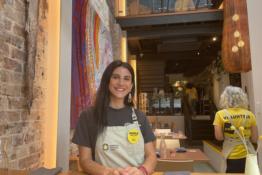 A young woman wearing an apron smiles inside a dimly lit restaurant