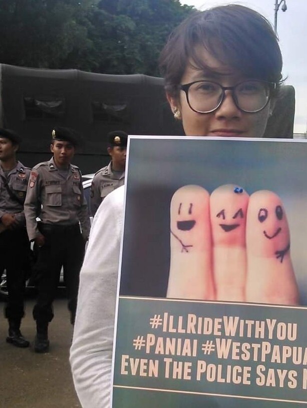 A woman holds a poster of three fingers with features drawn on them to look like people. Three policemen stand behind her.