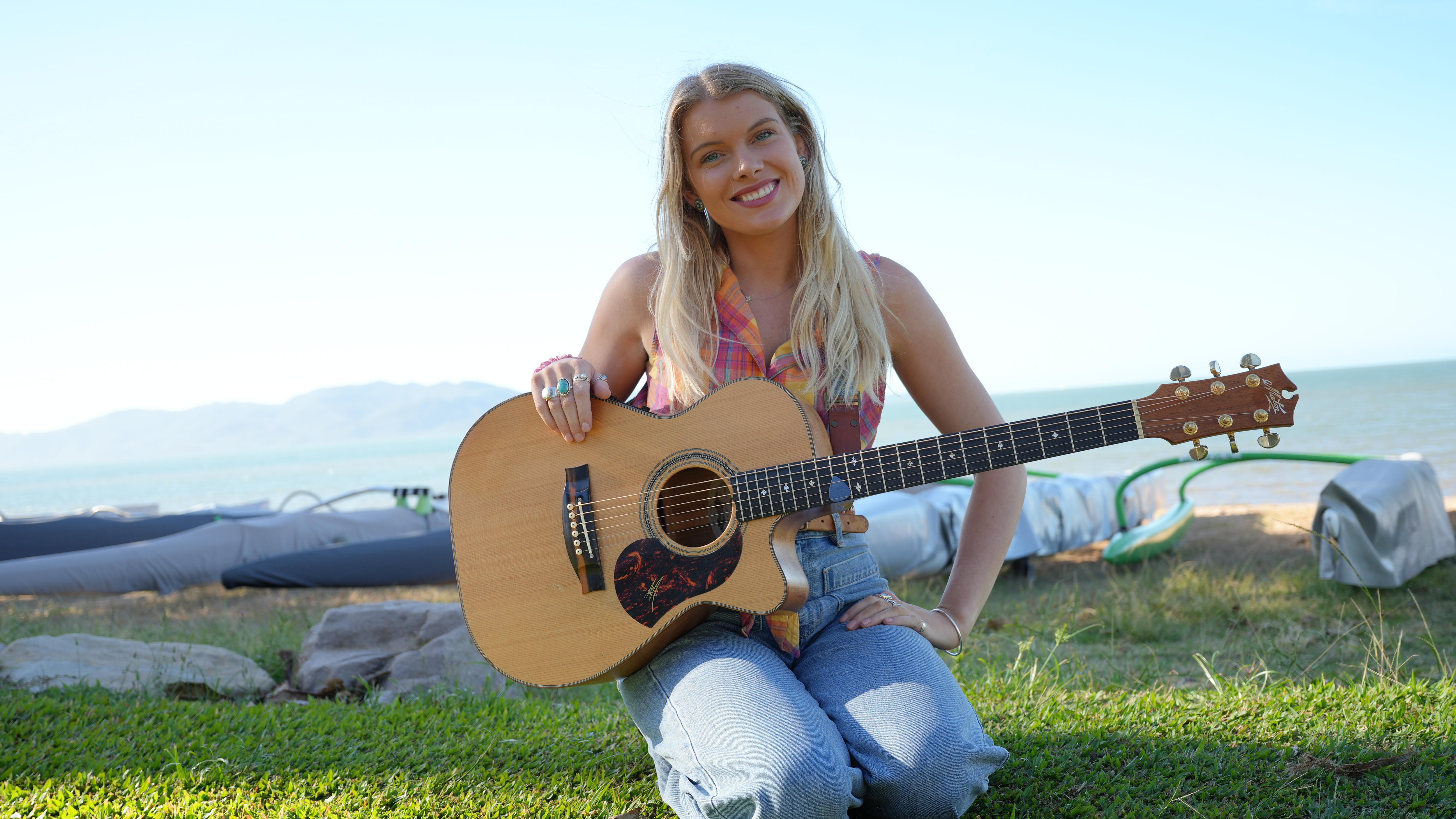 blonde woman with acoustic guitar sitting in front of beach