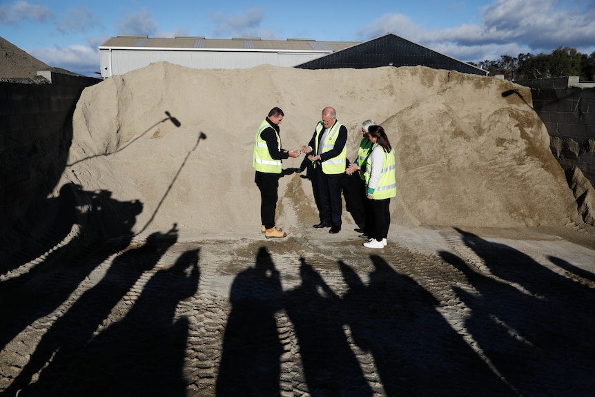 Morrison with people in high-vis vests looking at sand with shadows of media in foreground.