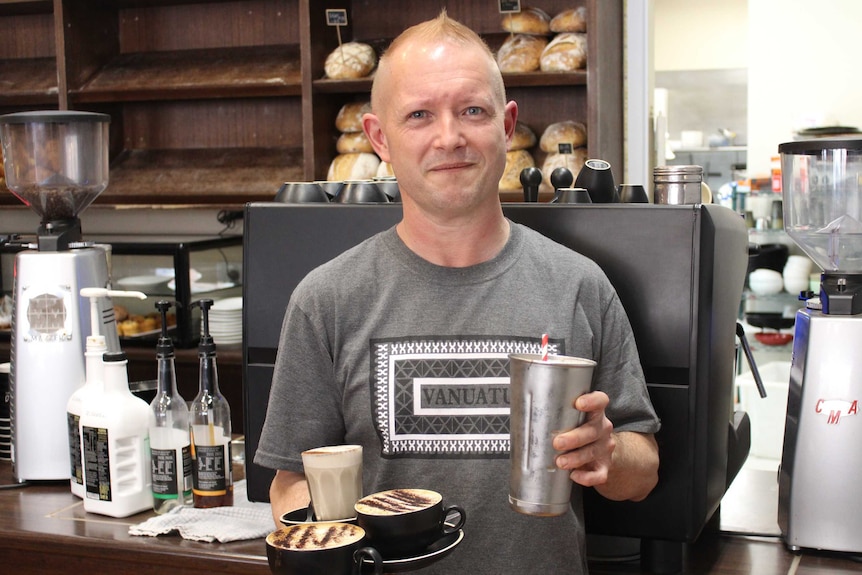 Mark standing in front of the coffee machine. He is holding three coffees and a milkshake.