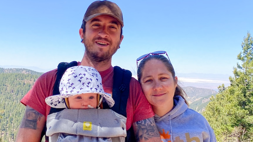 A man with a baby in a front carrier and a woman smile for a selfie 