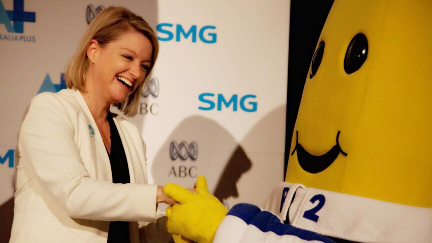 A woman smiles and holds hands with a banana in pyjamas in front of a promotional banner.