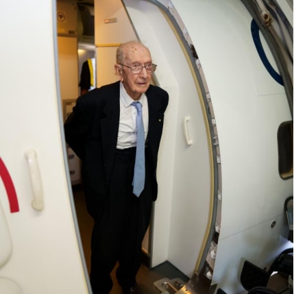 An elderly man wearing a suit and tie stands at the door to a large plane.